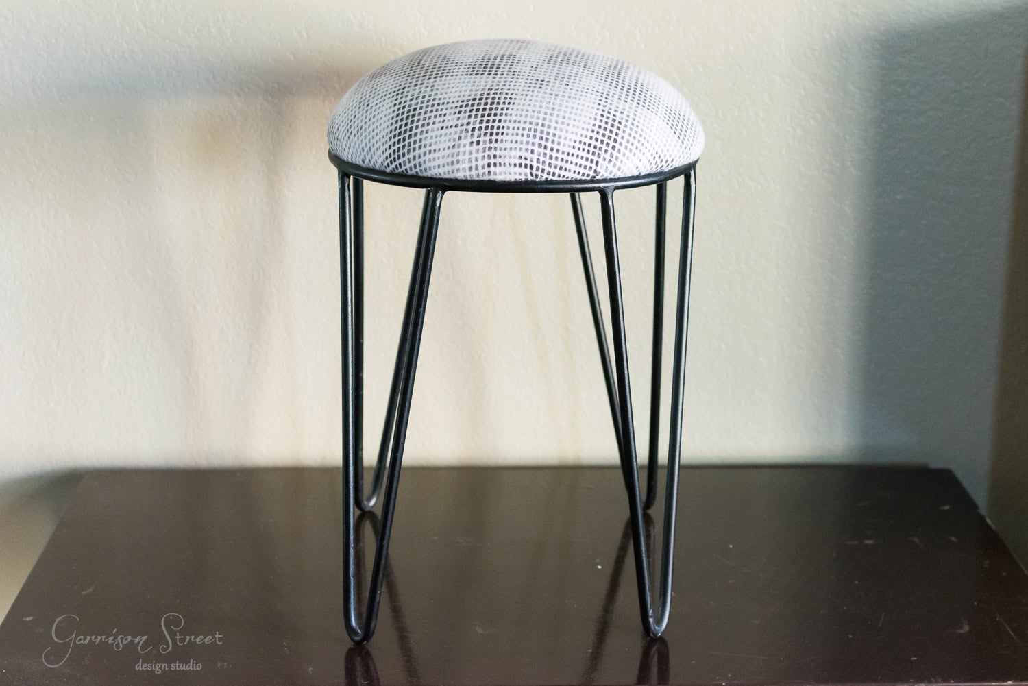 A small round stool with a vintage touch features a light gray cushioned top and black metal hairpin legs. It sits gracefully on a dark wooden surface against a light-colored wall, reminiscent of a well-crafted tool chest from bygone eras.