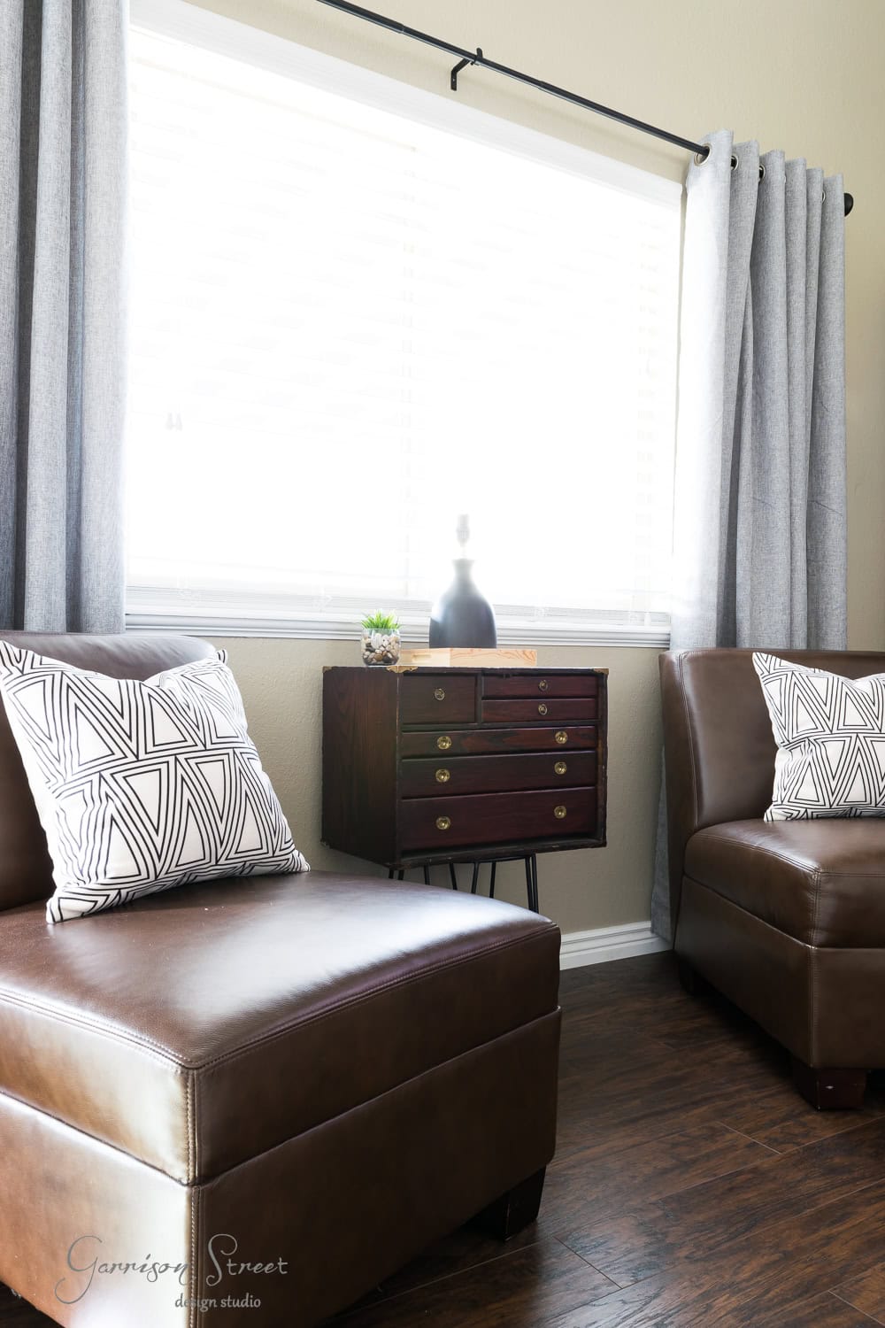 A cozy living room corner featuring two brown leather chairs with geometric patterned cushions. Between them is a vintage wooden side table with multiple drawers resembling a tool chest, adorned with decorative items. Grey curtains frame a window with blinds.