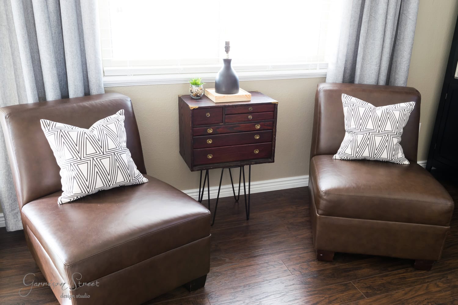 Two vintage brown leather chairs with geometric-patterned pillows face each other. A small wooden cabinet resembling a tool chest sits between them, topped with a lamp and plant. The room has a hardwood floor, and light enters through a window with curtains.