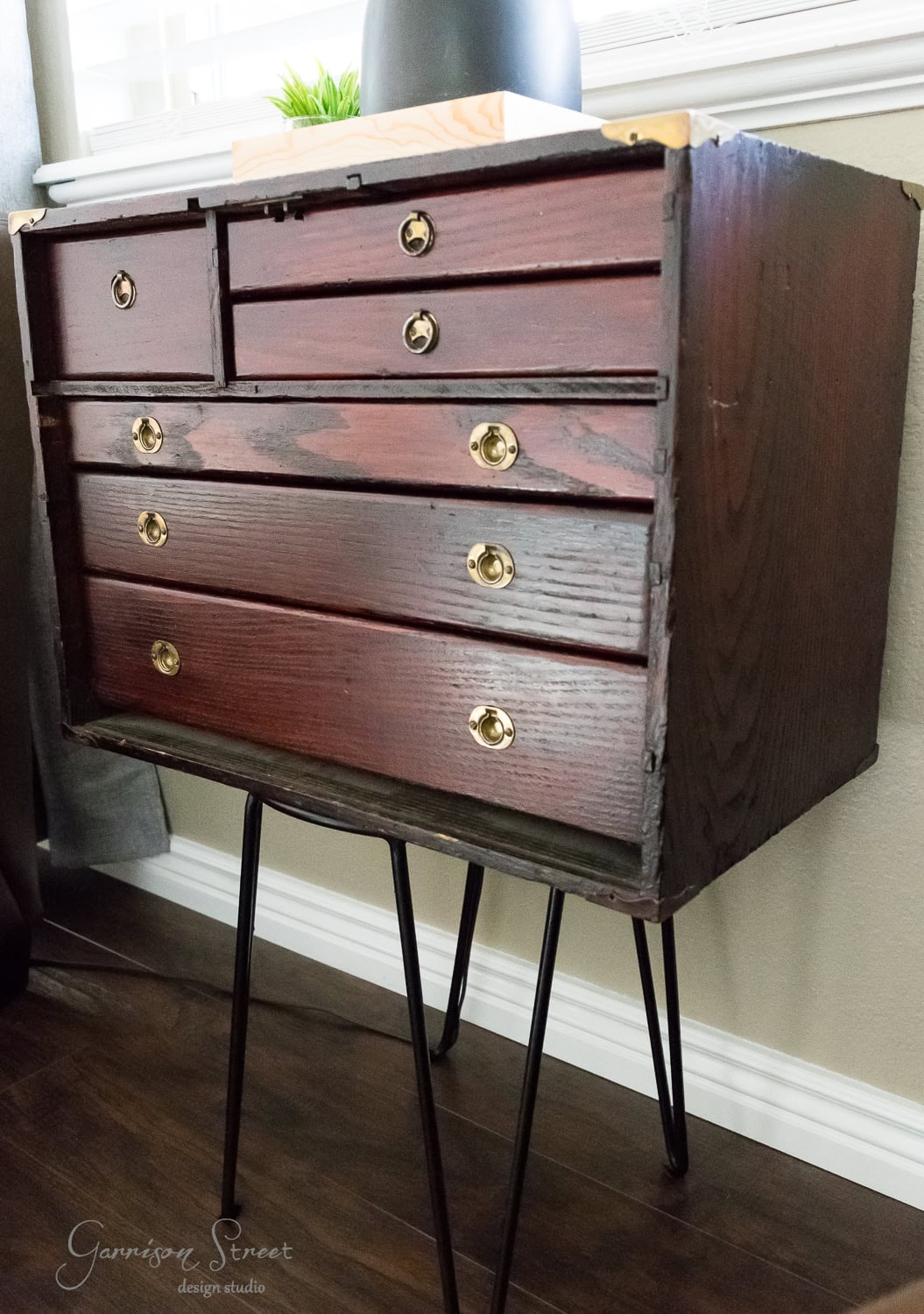 A vintage wooden dresser with six drawers, styled like a classic tool chest, features brass knobs. The dresser rests on black metal hairpin legs and is positioned in a well-lit room with a small plant on top.