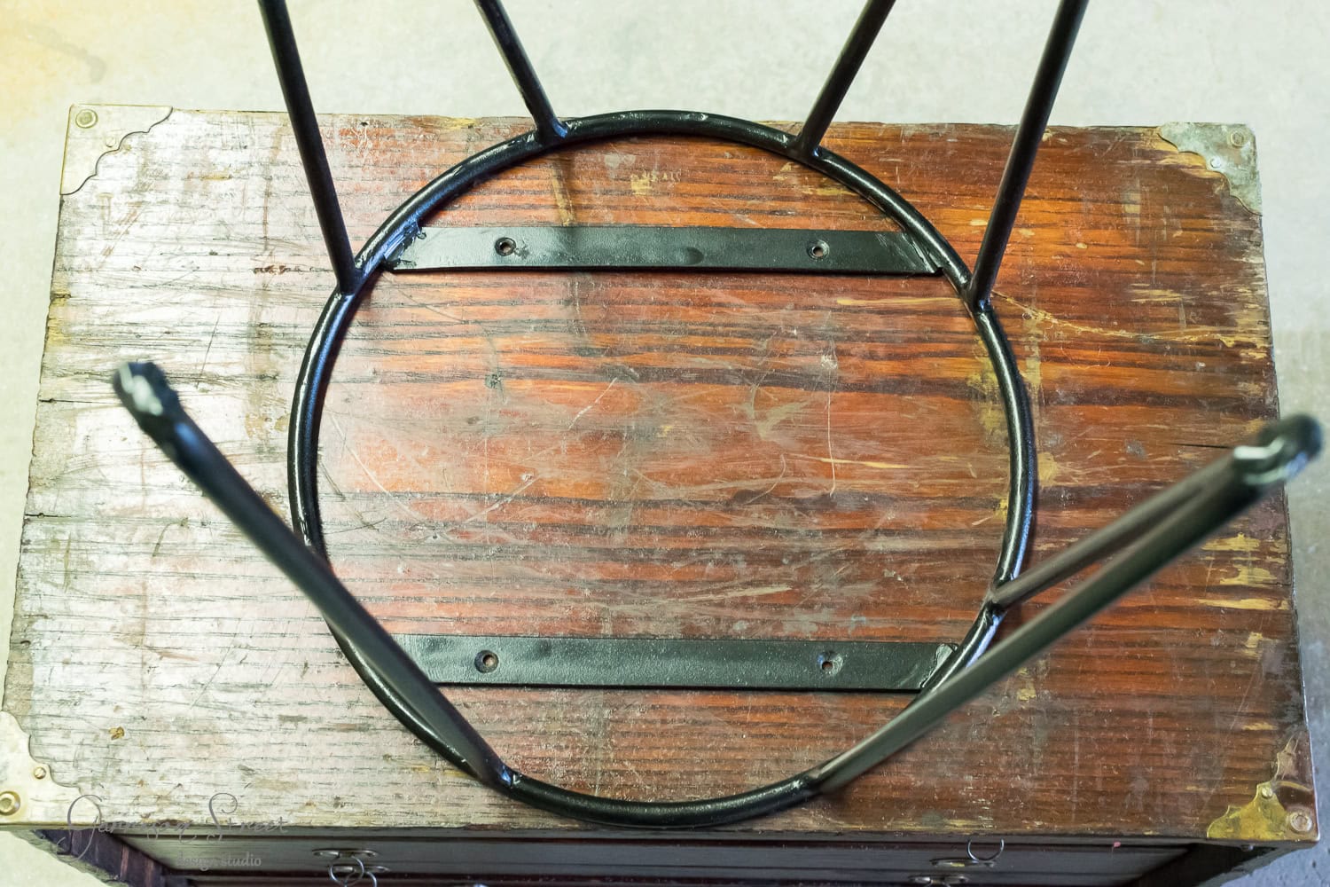 An upside-down stool with a circular metal frame and four legs rests on a vintage wooden surface. The wood has visible grain and wear, accented by small metal corner brackets, reminiscent of an old tool chest's sturdy craftsmanship.