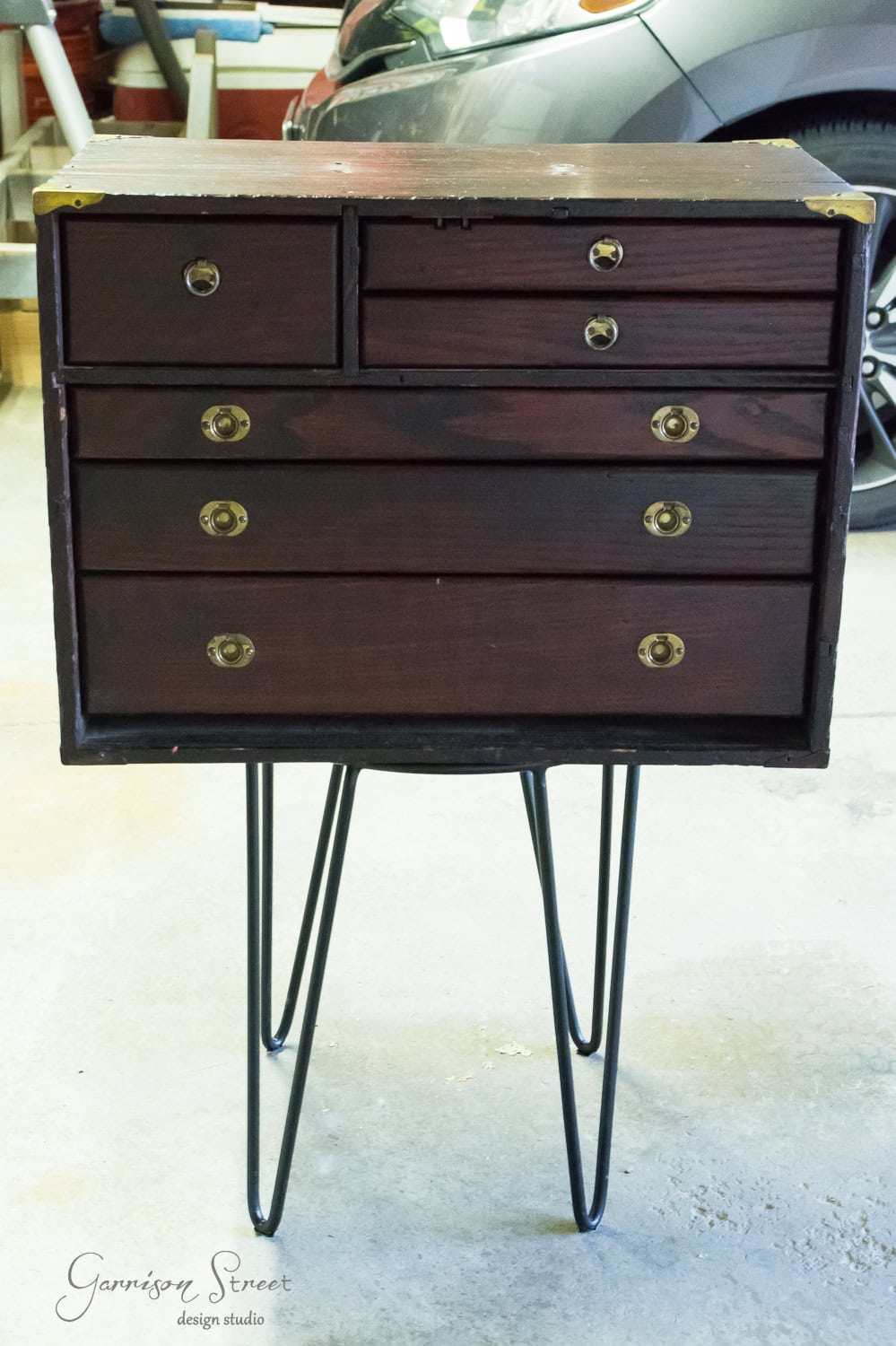 A small wooden dresser, reminiscent of a vintage tool chest, with four drawers and metal knobs, stands on sleek black metal hairpin legs. It is placed in the garage, with a car visible in the background.