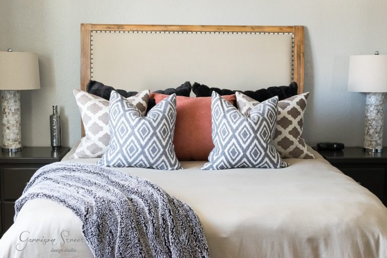 A neatly made bed with a beige headboard graces the guest room, adorned with patterned pillows in shades of grey, black, and pink. Two matching lamps sit on bedside tables, framing the bed perfectly. A textured throw is draped elegantly over the foot of the bed.