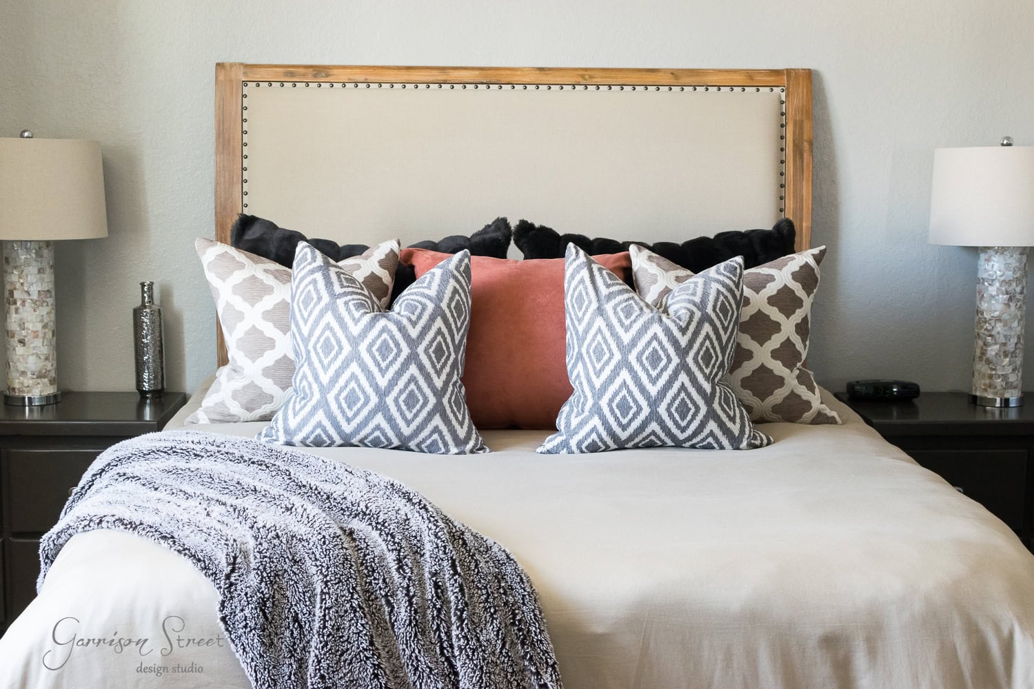 A neatly made bed with a beige headboard graces the guest room, adorned with patterned pillows in shades of grey, black, and pink. Two matching lamps sit on bedside tables, framing the bed perfectly. A textured throw is draped elegantly over the foot of the bed.
