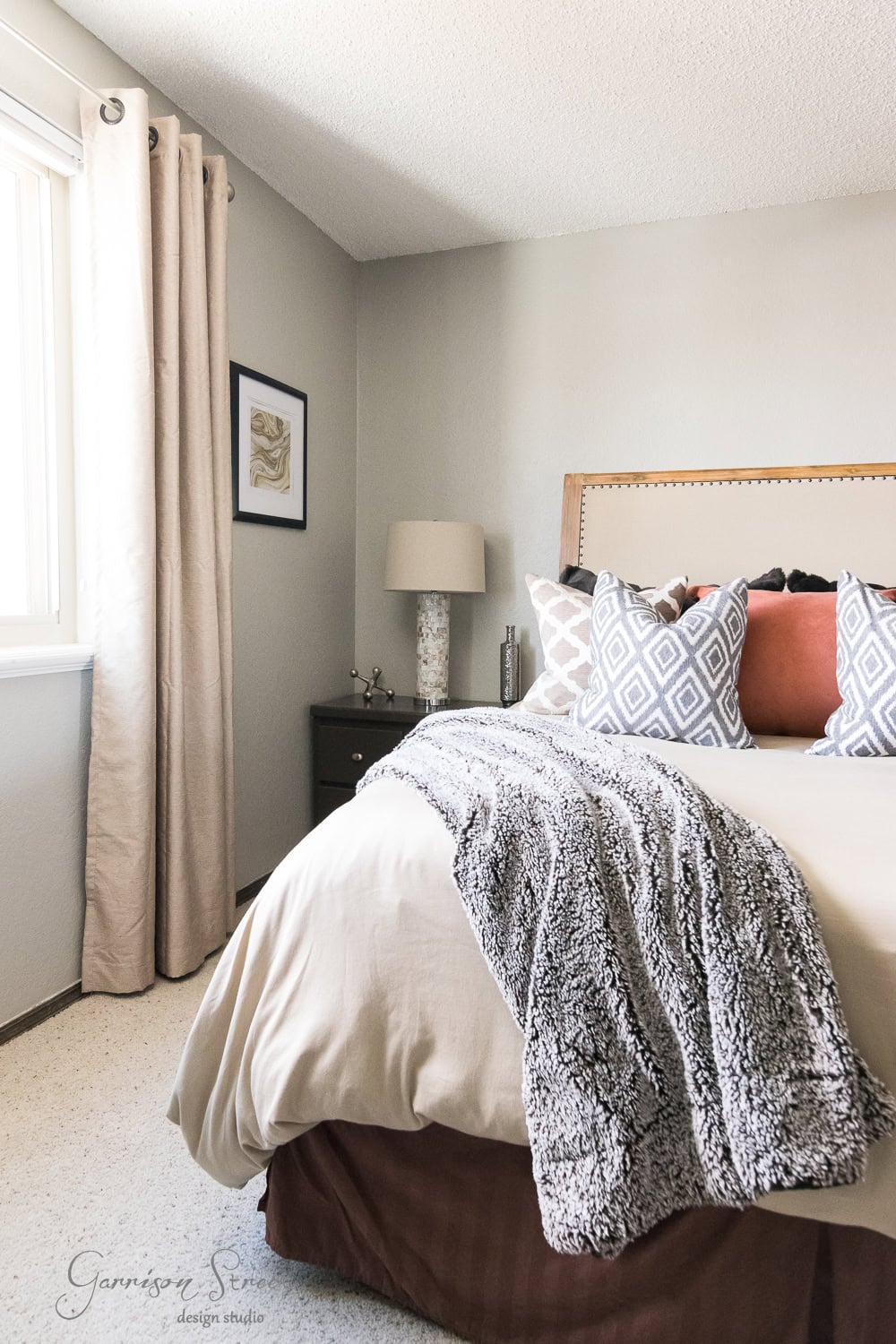 A cozy guest room features a large bed adorned with patterned pillows and a fuzzy blanket. A bedside table with a lamp sits nearby, while sunlight filters through the window's light curtains, casting gentle rays on the framed picture hanging on the wall.
