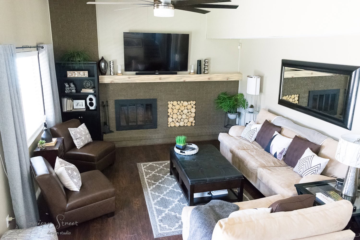 A cozy family room makeover featuring a beige sofa, dark coffee table, and armchairs. The space has a wall-mounted TV above a fireplace with wood accents, a large mirror, and a ceiling fan. Decorative plants and a patterned rug grace the wooden floor.