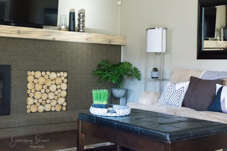 A cozy living room features a beige sofa with patterned pillows, a dark wood coffee table, and a fireplace decorated with stacked logs. A floor lamp and green plants add warmth, while a mirror and TV adorn the light gray walls.