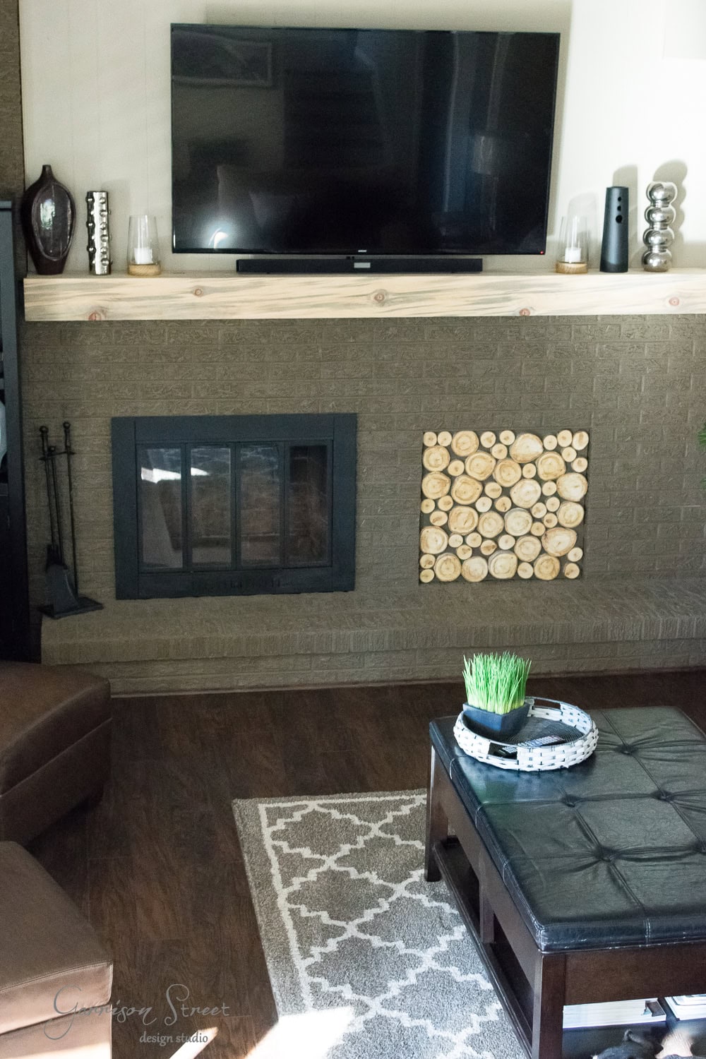 A cozy family room features a mounted TV above a fireplace framed by decorative wood slices. The dark wood coffee table with a plant and tray sits on a patterned rug, adding charm to the room's earthy, modern look—a recent makeover that highlights neutral-toned decor and seating.