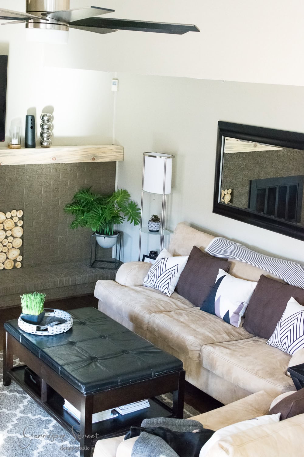 A cozy family room features a beige sofa, dark cushions, and a black coffee table. A geometric rug covers the floor, while a fireplace with stacked logs and a wooden mantel sits on the left. A large mirror and plants enhance the decor, completing this stylish makeover beneath a ceiling fan.