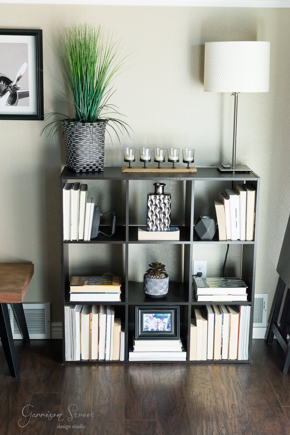 Bookshelf makeover in the family room featuring a variety of books, a black and white vase, and decorative candles. A potted plant and lamp sit on top. A small framed picture and a decorative item are nestled within the shelves. The dark wood floor completes the cozy ambiance.