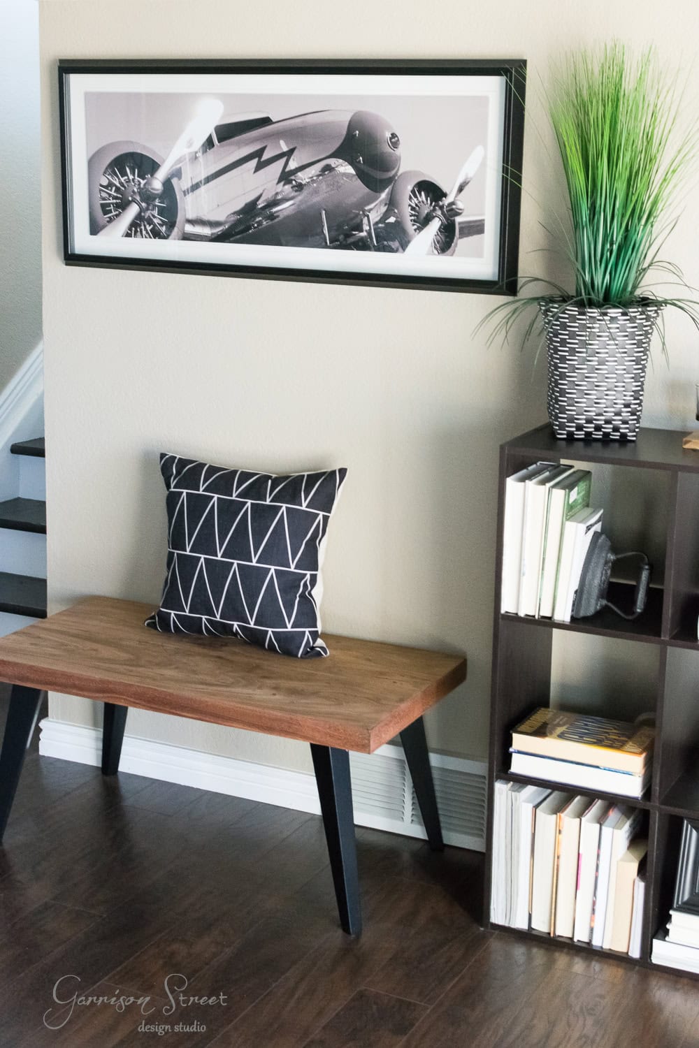 A modern family room makeover showcases a wooden bench with a black and white geometric pillow, complemented by a framed airplane photo on the wall above. A dark bookshelf filled with books and a potted plant enhances the polished wood floor's elegance.