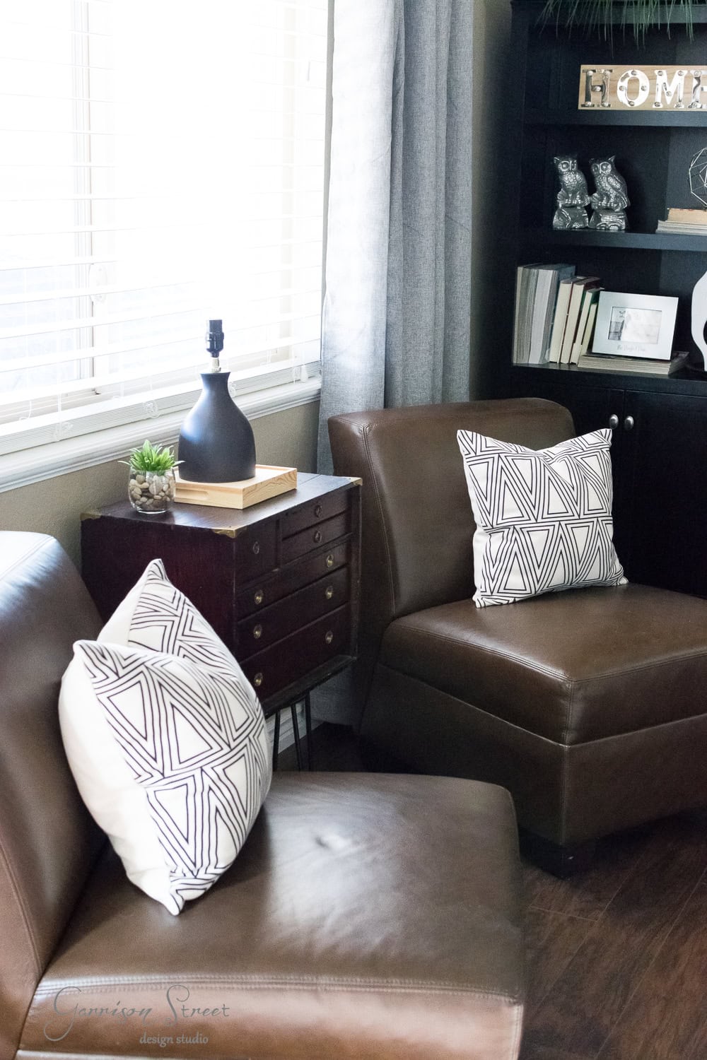 A cozy family room featuring two brown leather chairs with geometric-patterned cushions. A small wooden cabinet, topped with a black lamp and a plant, sits between them. Books and decor items fill a dark bookshelf in the corner, giving the space a stylish makeover.