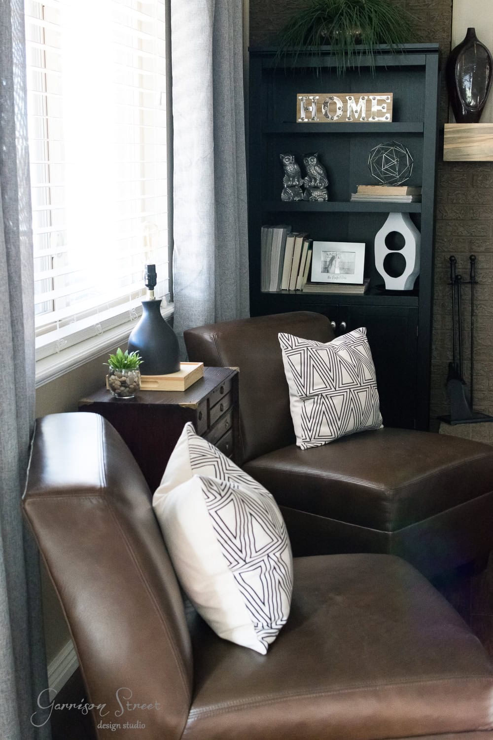 A cozy family room corner with two brown leather chairs featuring geometric-patterned pillows. A black bookshelf showcases books, decorative items, and a framed photo. A large window with curtains lets in natural light. After a recent makeover, a small side table now holds a stylish lamp.