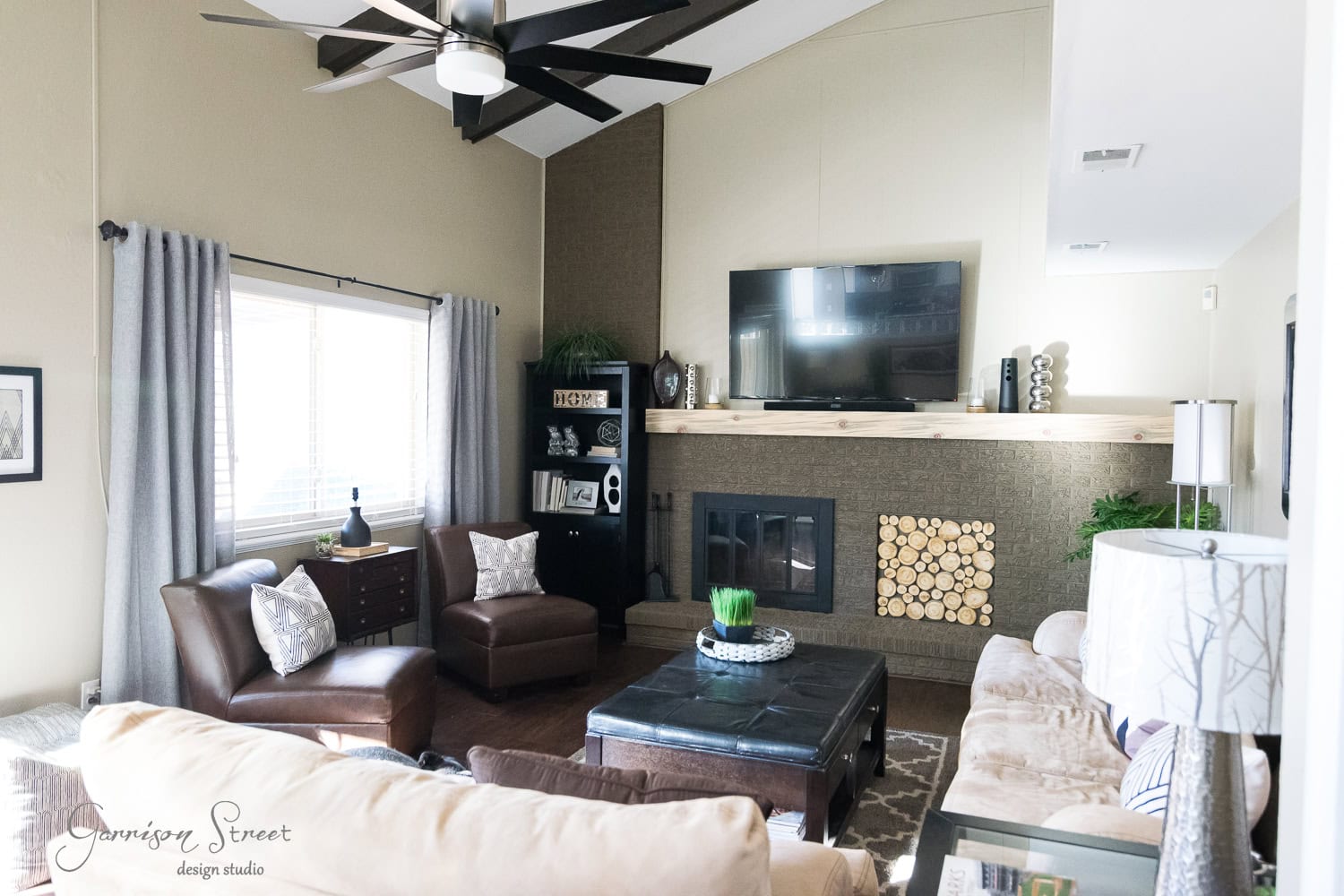 A cozy family room featuring a brown leather couch and armchairs, a dark wooden coffee table, a fireplace with wood storage, and a large TV mounted above the mantel. A recent makeover added a ceiling fan, gray curtains by the window, and decorative plants alongside displayed books.