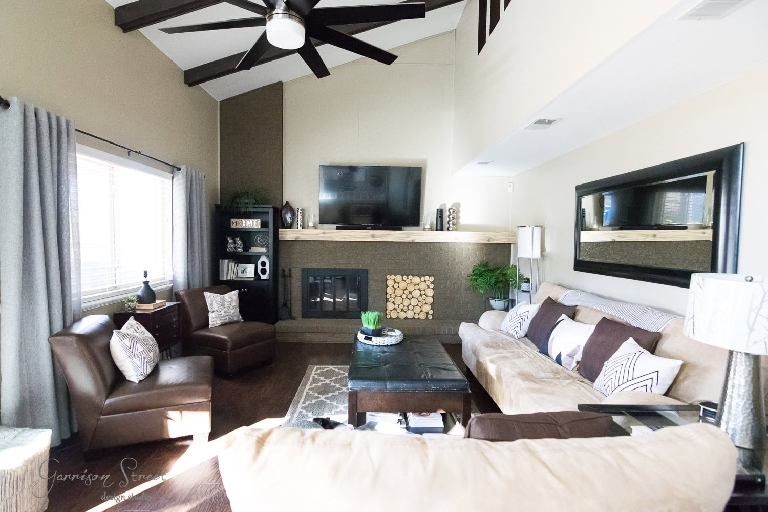A spacious family room with vaulted ceilings and a ceiling fan. It features a large beige sectional sofa, a coffee table, and two brown armchairs. A fireplace is set into a wall with a TV above it. Natural light streams in through large windows, completing the perfect home makeover.