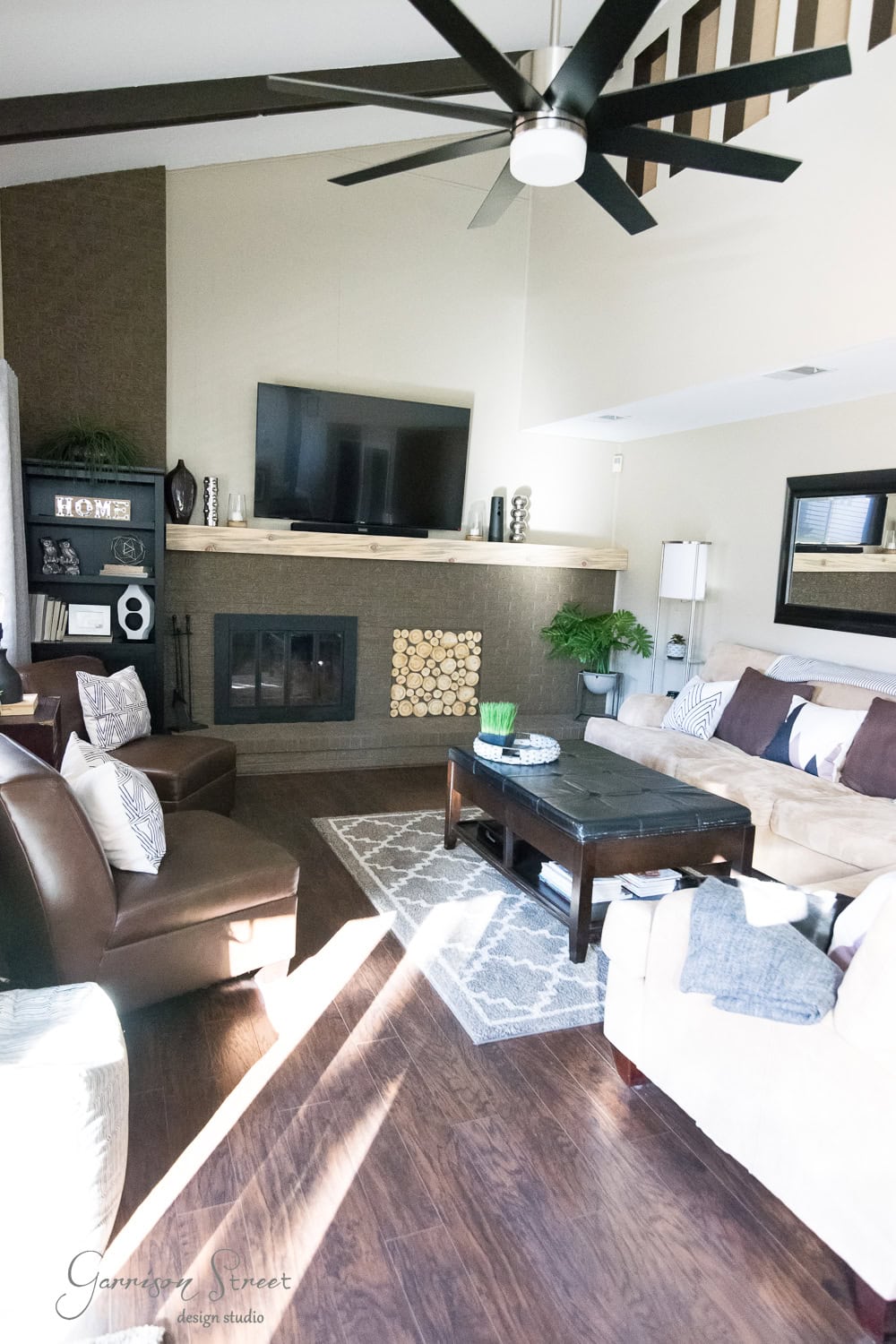 A cozy living room with a beige sofa, wooden coffee table, and armchairs. A large TV is mounted above a fireplace with a wooden mantel. Decorative shelves and a patterned rug complete the space, bathed in natural light.