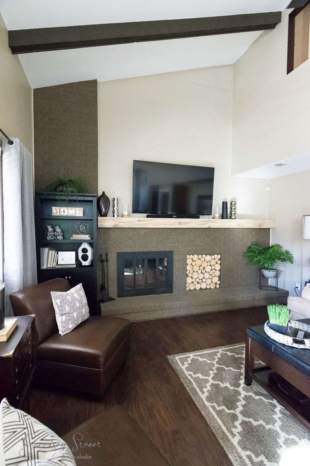The family room boasts a modern makeover with a mounted TV above the stylish fireplace. A dark brown armchair with a geometric pillow sits on the left, complemented by wooden floors, a patterned rug, potted plants, and decorative shelves adorned with books and ornaments.