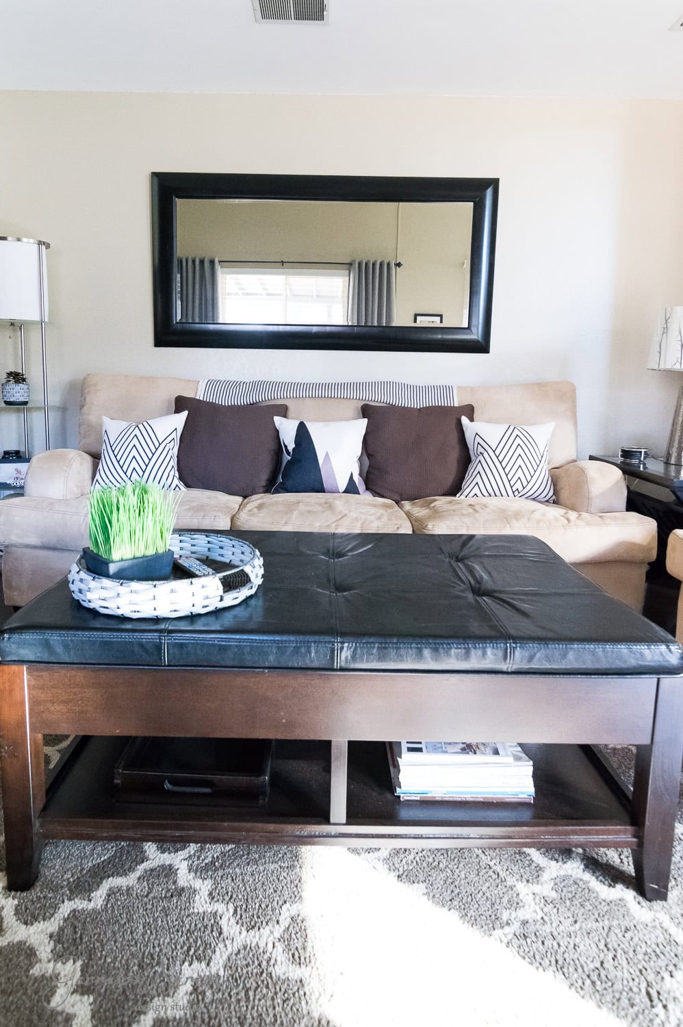 A cozy family room features a beige couch with patterned pillows, a large black-framed mirror above, and a dark wood coffee table with a black cushion top. The table holds a decorative grass plant and round tray. Neutral tones and sunlight create a warm atmosphere, offering the perfect makeover touch.