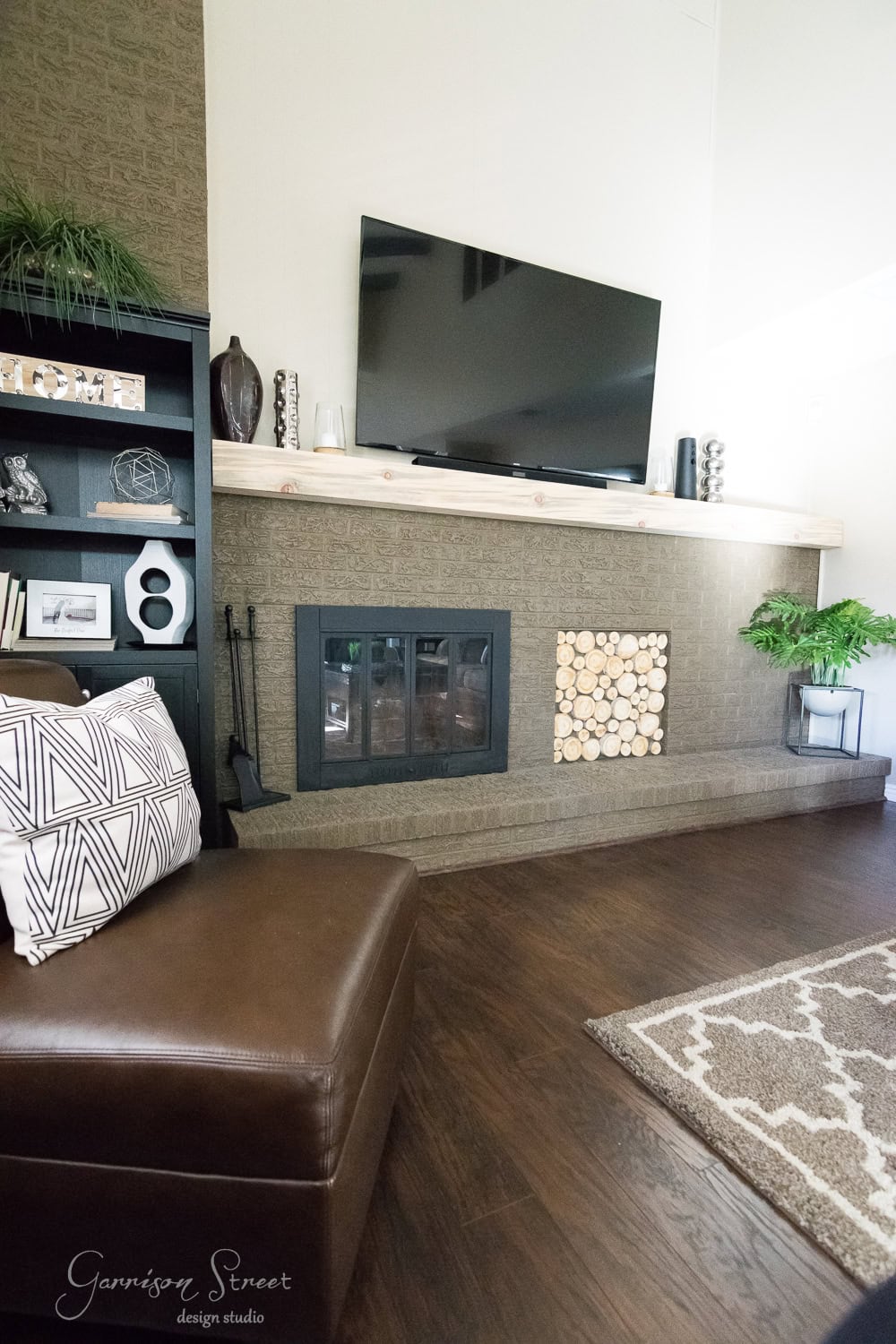 Experience a stunning family room makeover featuring a modern fireplace with a flat-screen TV above it and decorative wood slices beside. A brown leather chair with a geometric pillow, along with a dark shelf of decor, complement the leafy plant on the wooden floor beautifully.