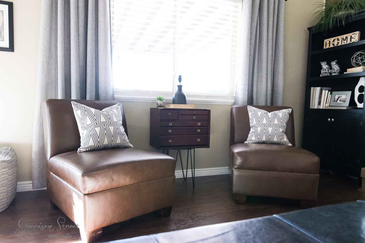 A cozy family room showcases a pair of brown leather chairs adorned with geometric-patterned pillows in front of a window framed by gray curtains. Between the chairs is a small wooden chest, hinting at its recent makeover, while a black shelf with decor stands gracefully on the right.