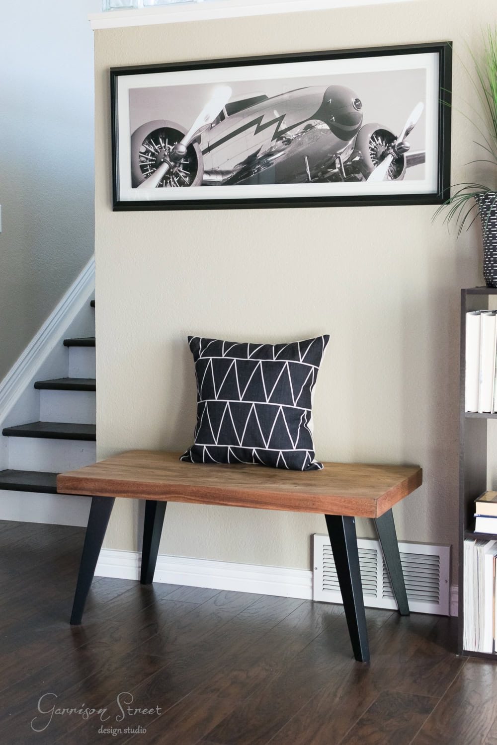 A modern entryway features a wooden bench with black legs, perfectly blending into the recent makeover. A geometric-patterned pillow rests on the bench. Above, a framed black-and-white photo of a vintage airplane hangs on a beige wall. A staircase leading to the family room is visible on the left.