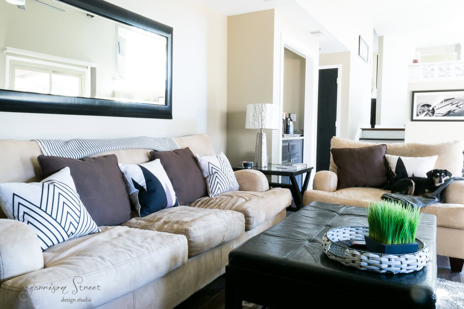 A modern family room makeover features a beige couch with brown cushions, a black coffee table adorned by a plant, and a mirror on the wall. A dog rests on a beige armchair as natural light filters through, creating a cozy atmosphere.