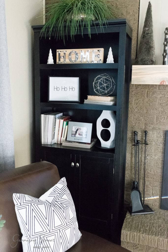 Free Printables A dark wood bookshelf with the word "HOME" on the top shelf, surrounded by Christmas decorations like books, a framed "Ho Ho Ho" sign, and geometric decor. A brown leather armchair with a patterned pillow is positioned in front, adding a modern touch to the cozy setting.