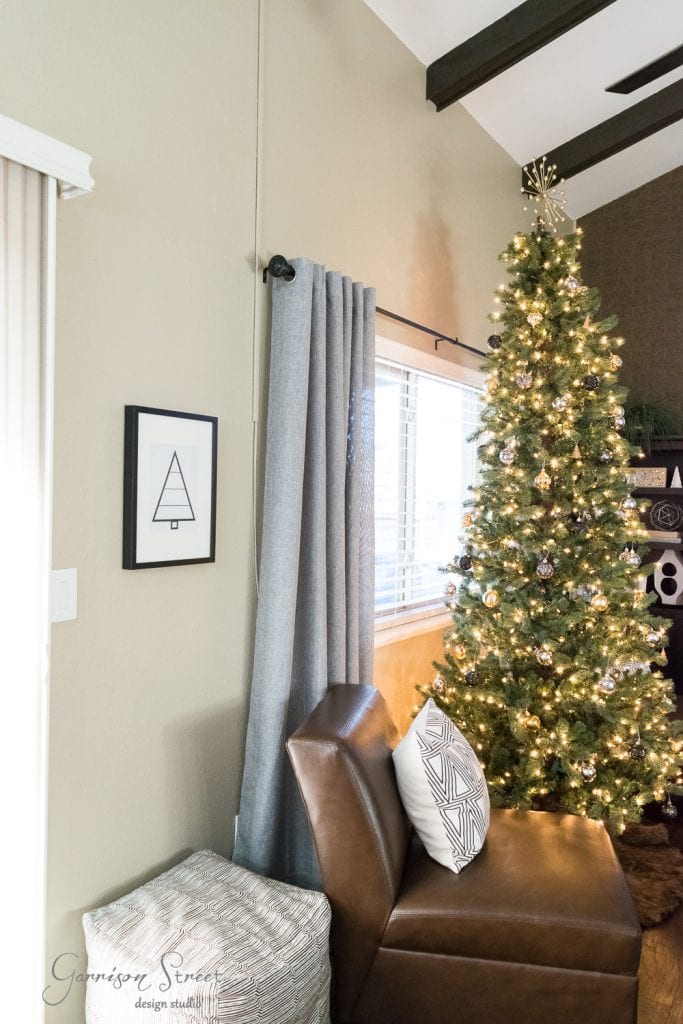 Free Printables A modern living room features a tall, decorated Christmas tree with lights near a window. A brown leather chair and geometric-patterned pillow rest beside a textured ottoman. Framed minimalist tree artwork and festive printables add charm to the beige wall.