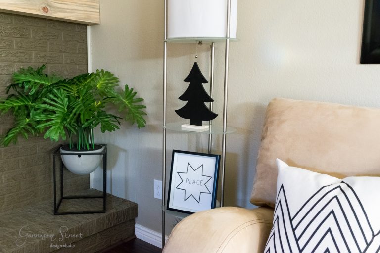 A cozy living room corner featuring a beige couch with a geometric-patterned pillow. There's a small tall shelf with a lamp, a black tree decoration, and a "Peace" framed print. A large green plant sits beside a brick fireplace.