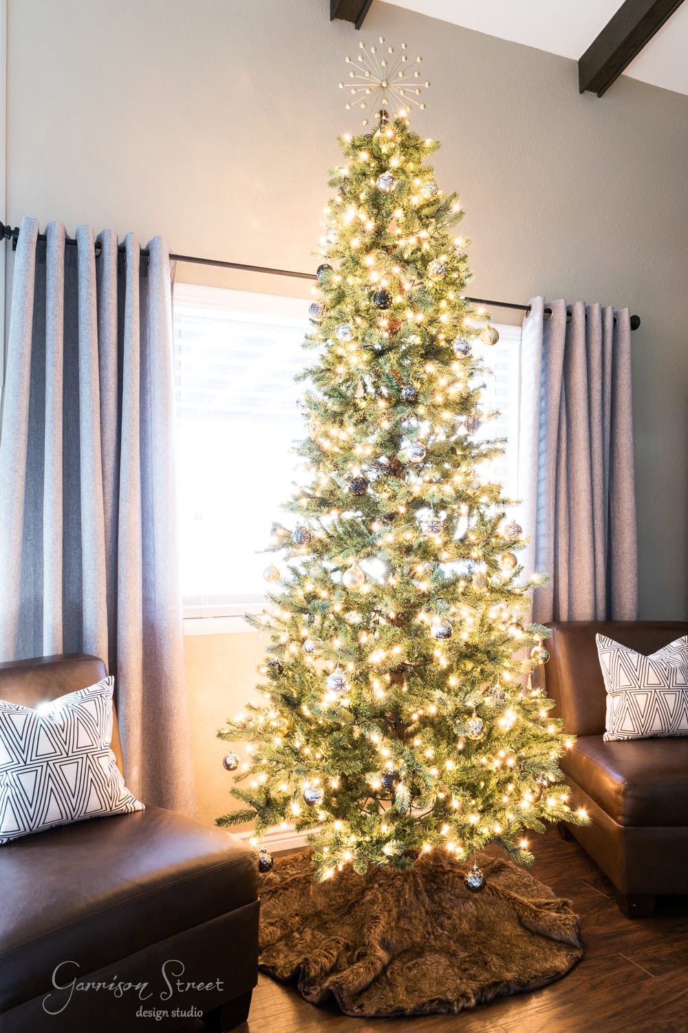 A tall, slender Christmas tree adorned with white lights and shiny ornaments stands between two brown chairs with patterned cushions. The room exudes a modern charm, softly lit with gray curtains framing a window in the background.
