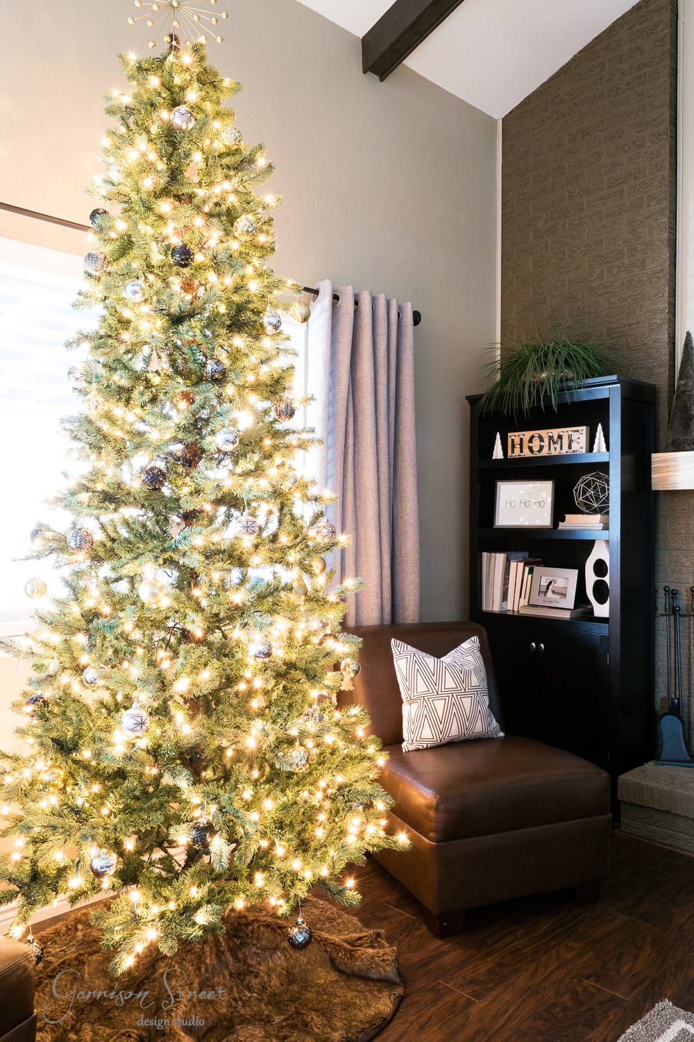 A modern, decorated Christmas tree with white and silver ornaments stands in a cozy living room. Next to it is a brown armchair with a geometric-patterned cushion, and a bookshelf with decorative items subtly complements the festive scene.