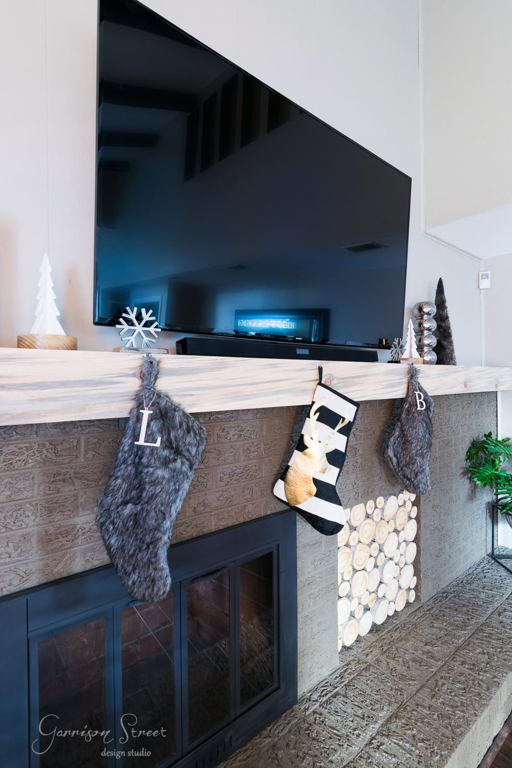 A fireplace mantel adorned with two gray stockings labeled "L" and "B," a modern Christmas tree ornament, and a large flat-screen TV above. The hearth showcases stacked wood decor, enhancing the contemporary aesthetic.