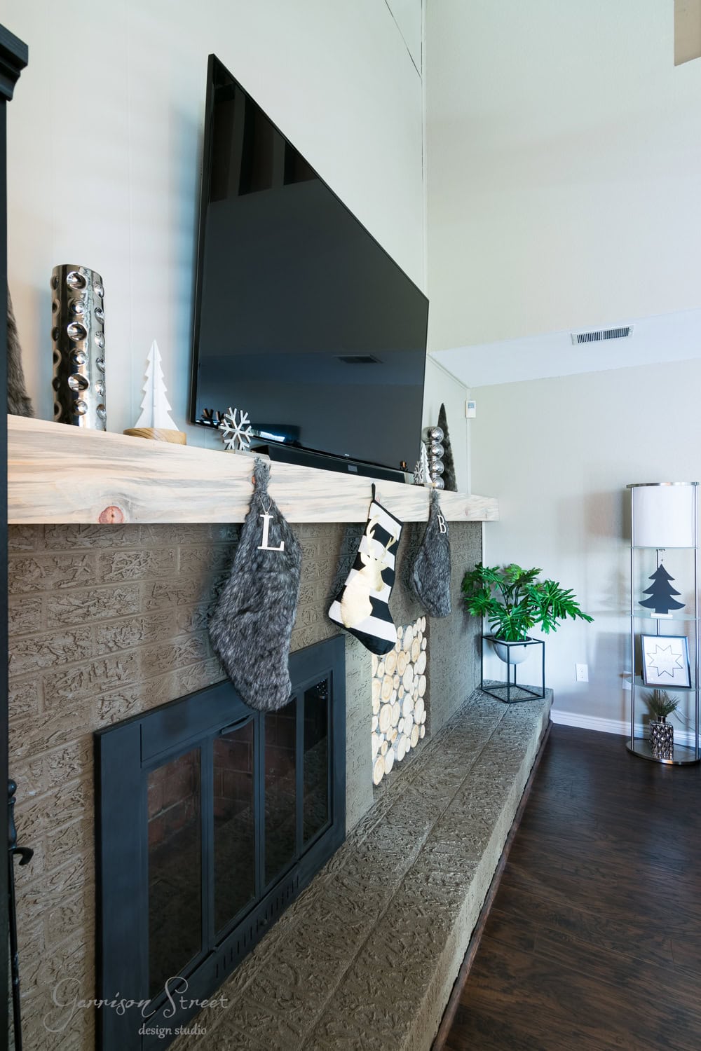 A cozy living room with a brick fireplace elegantly decorated for Christmas. Three stockings hang on the mantel beneath a mounted TV. A stack of firewood and a potted plant are nearby, adding warmth to the modern, minimalist decor.