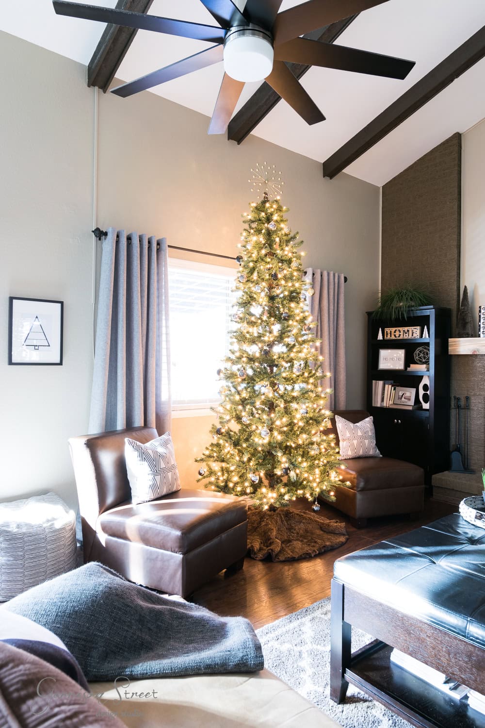 A cozy, modern living room with a tall Christmas tree lit by white lights. It features brown leather chairs and a sleek black bookshelf adorned with decorative items. A ceiling fan hums gently as light filters through closed curtains, casting a warm, inviting glow.