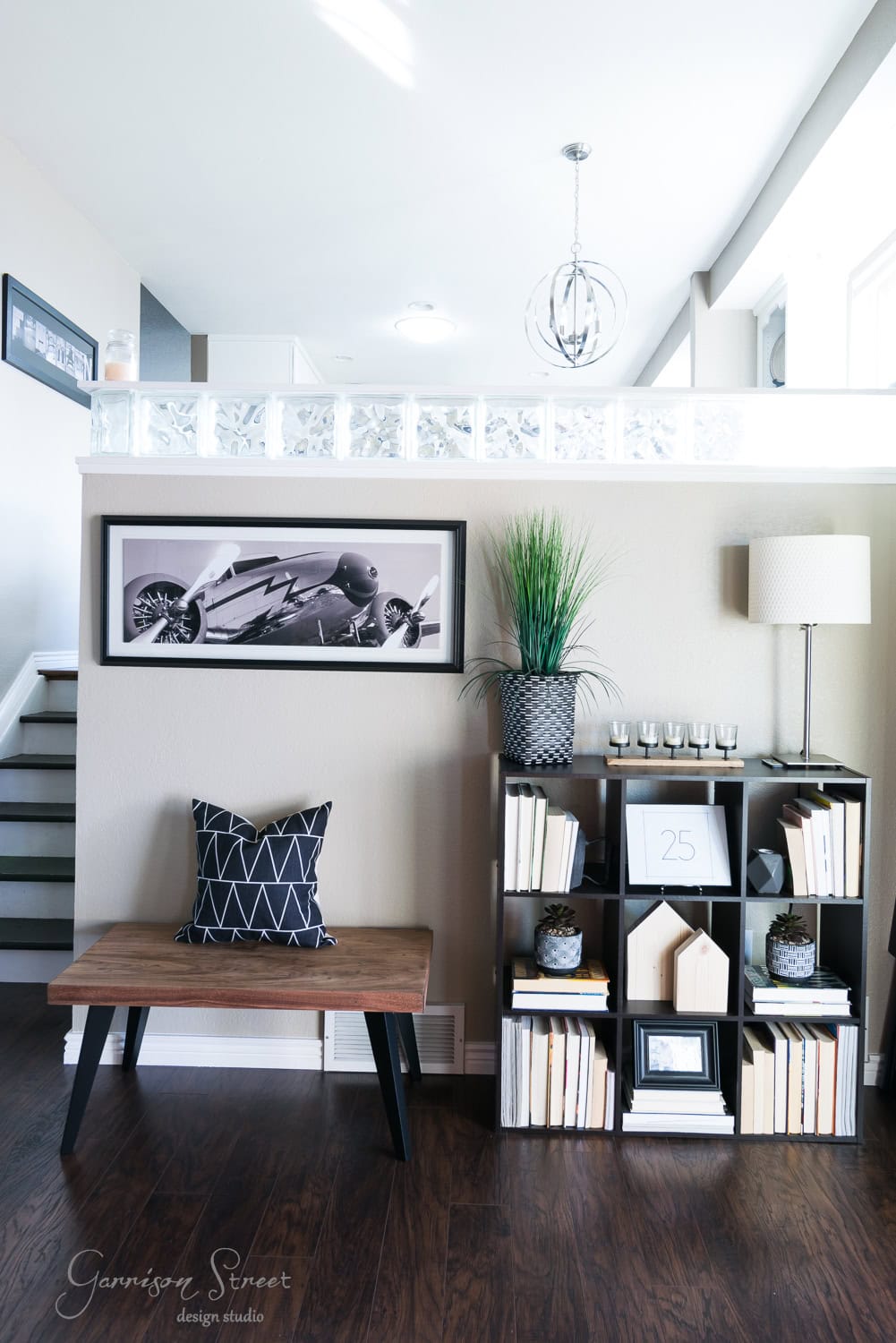 A modern living room with a wood bench and a black geometric pillow is adorned with festive Christmas touches. A framed motorcycle picture hangs above, while the bookshelf holds books, candles, and holiday decor, including a potted plant and lamp. Sunlight streams in, enhancing the room's airy feel.