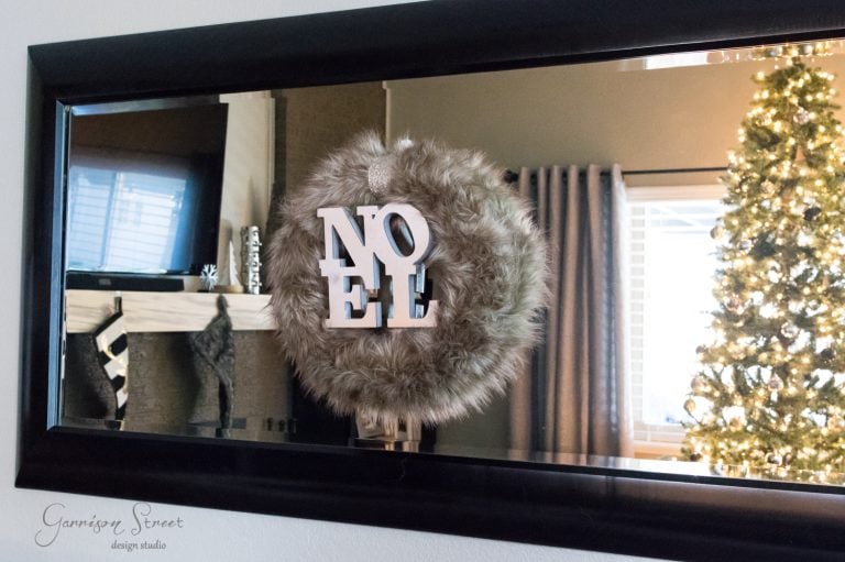 A fluffy wreath with the word "Noel" hangs on a mirror, reflecting a living room with a decorated Christmas tree and a fireplace mantel.