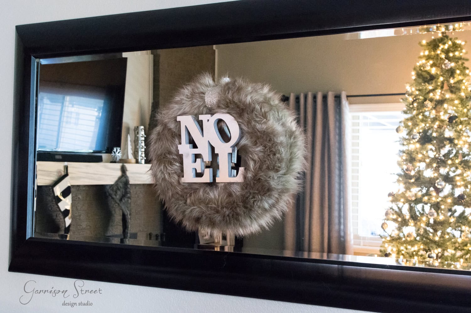 A fluffy wreath with the word "Noel" hangs on a mirror, reflecting a living room with a decorated Christmas tree and a fireplace mantel.