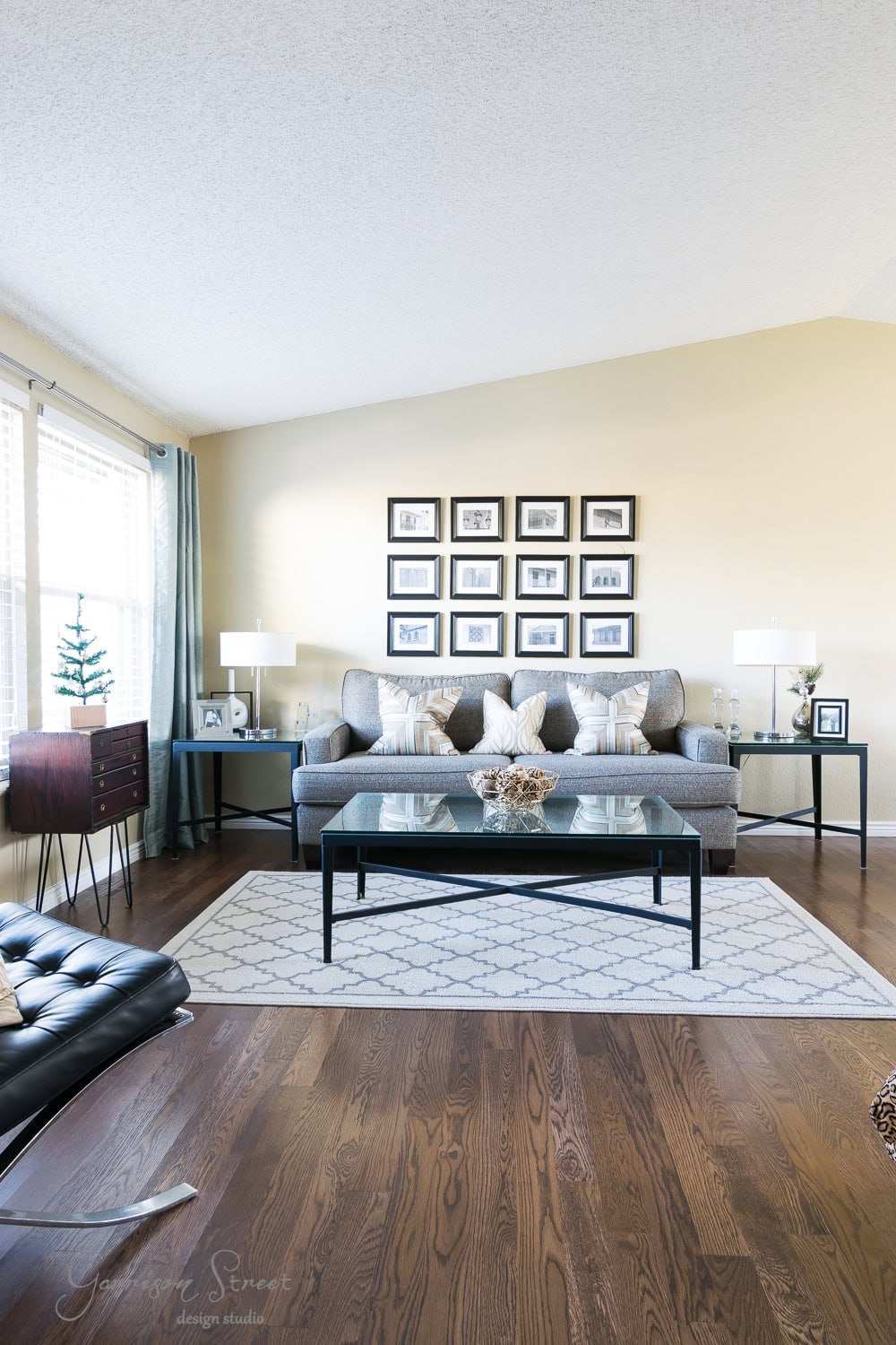 A spacious, modern living room with a beige sofa, patterned cushions, and a glass coffee table on a white geometric rug. The back wall features a grid of framed photos. Wooden flooring and large windows create a bright, inviting atmosphere perfect for cozy Christmas gatherings.