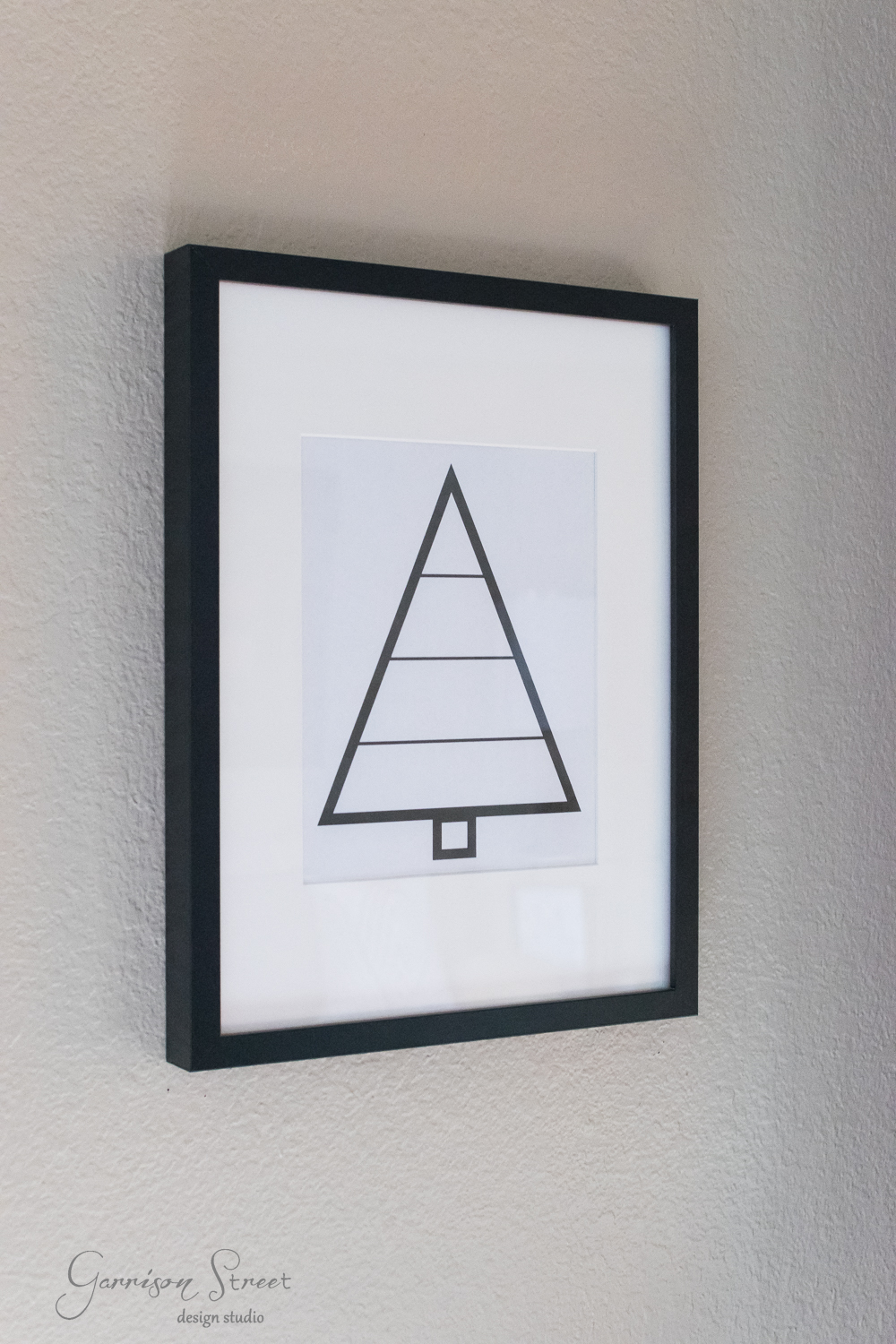 A framed minimalist artwork hangs on a wall, featuring a modern geometric design of a triangle resembling a Christmas tree with horizontal lines and a square at the bottom. The black frame offers striking contrast against the light-colored wall.
