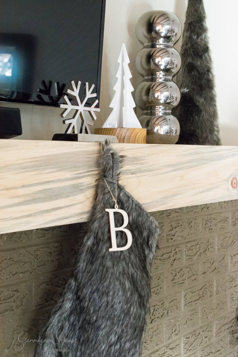 A modern holiday-themed mantel features a fluffy gray stocking with the letter "B," complemented by a wooden snowflake, a white tree, stacked silver ornaments, and a furry tree shape. This Christmas display brings together contemporary elegance and festive cheer.