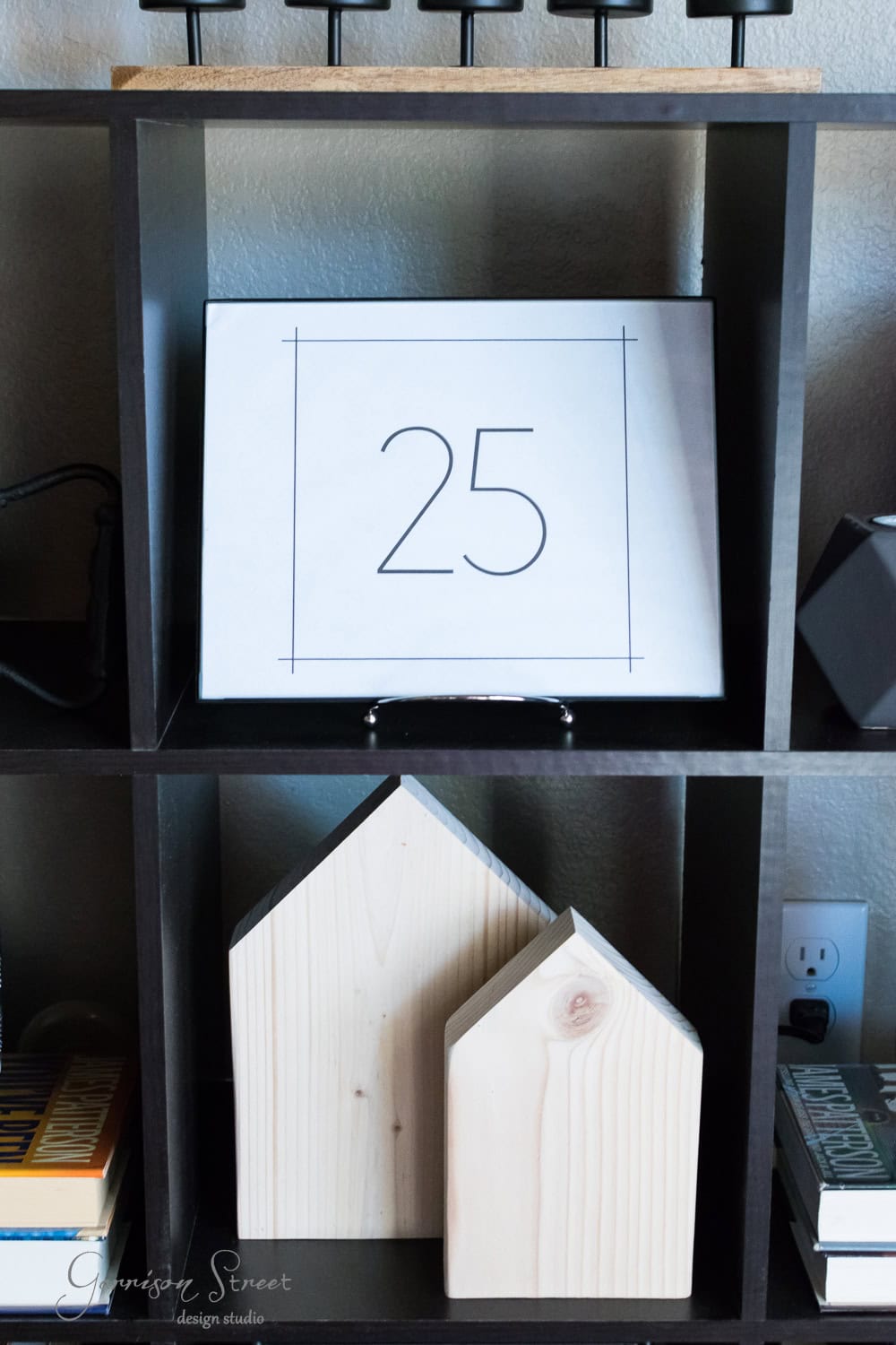 A numbered paper reading "25" is placed on a shelf, lending a subtle nod to Christmas. Below it are two modern wooden house-shaped decorations. Nearby, books and other decorative items add charm. The backdrop includes a wall socket and various sleek shelf compartments.