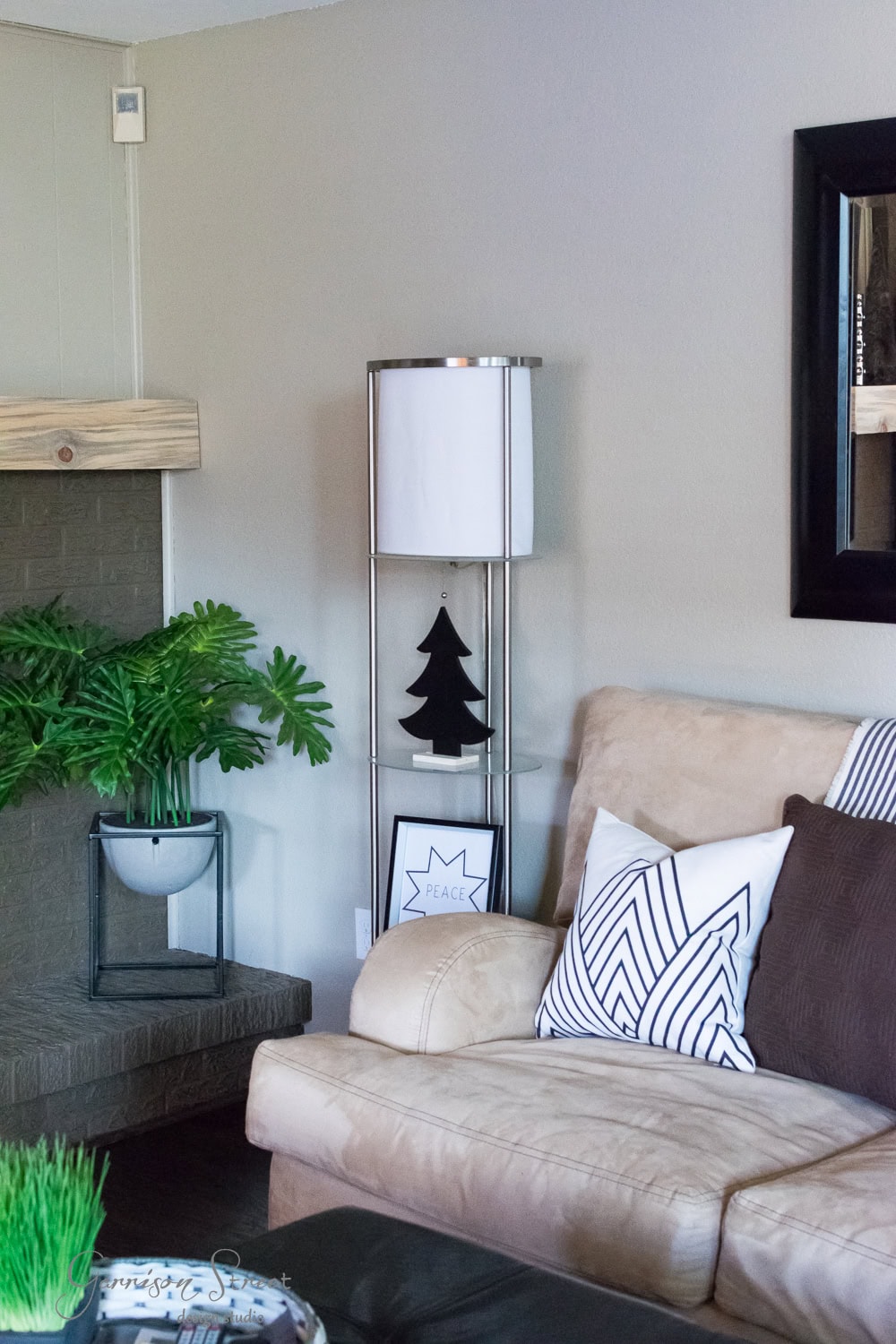 A cozy, modern living room features a beige couch with patterned pillows. A tall lamp and decorative tree silhouette sit on a side table, hinting at Christmas charm. A leafy plant graces the mantel, while a framed picture hangs on the wall. Neutral tones create a calm atmosphere.