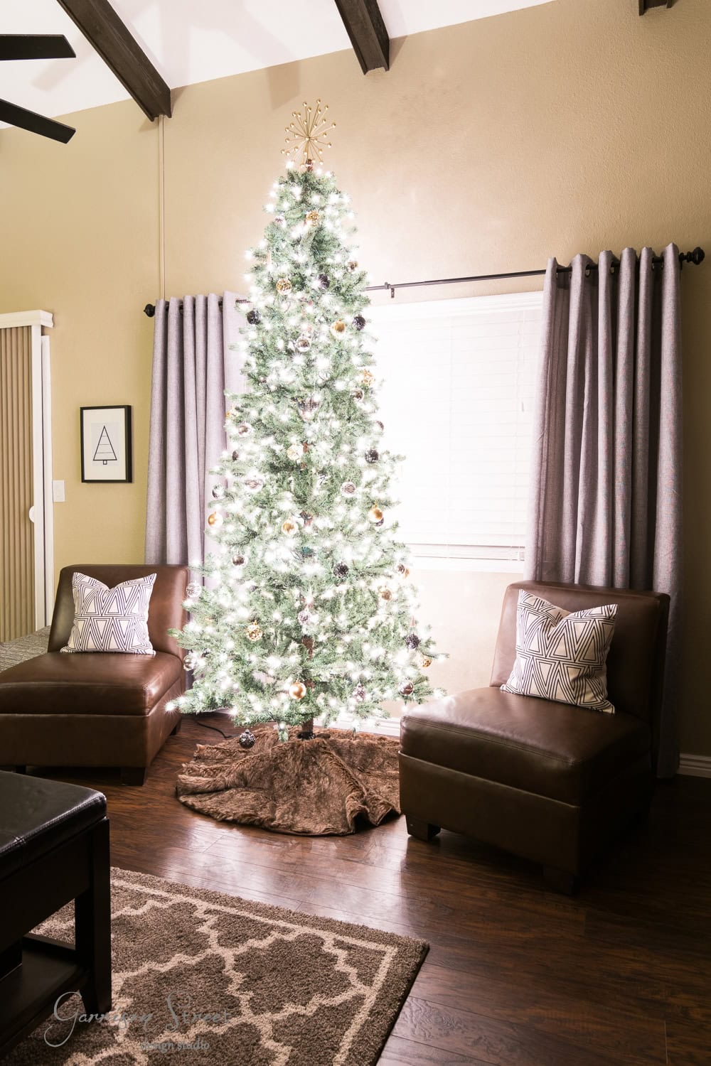A decorated Christmas tree with white lights and ornaments stands between two brown chairs in a cozy, modern living room. The room features wooden floors, a patterned rug, and a window with sleek gray curtains.