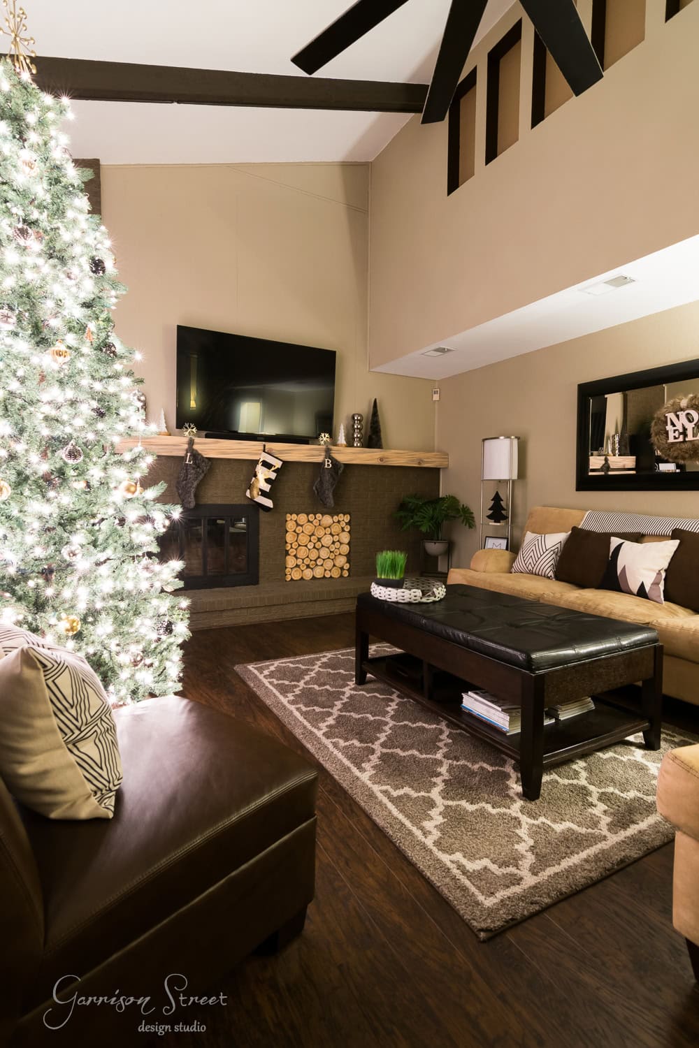 A cozy, modern living room showcases a beautifully decorated Christmas tree beside a fireplace adorned with stockings. The space features a sleek sectional sofa, a wooden coffee table on a patterned rug, and a flat-screen TV seamlessly integrated above the fireplace.
