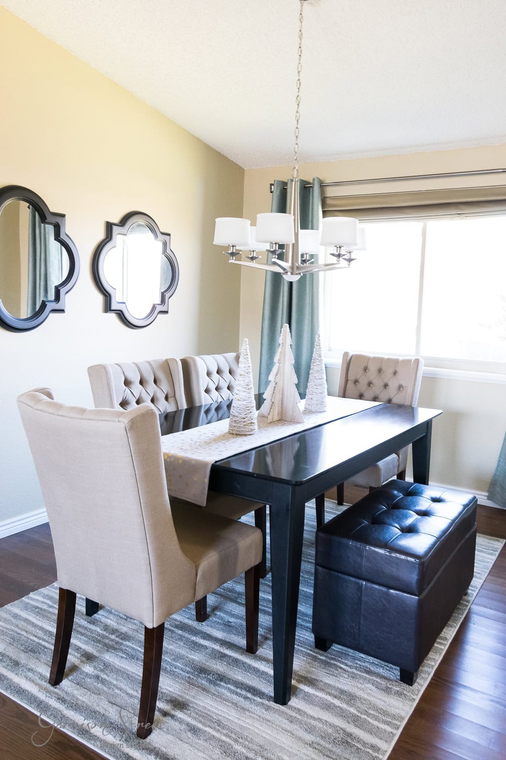 A modern dining room showcases a dark wooden table with a tufted bench and chairs. Festive cone-shaped trees add a touch of Christmas cheer atop the table. A chandelier hangs above, while two mirrors adorn the beige wall. A large window with curtains brightens the room.