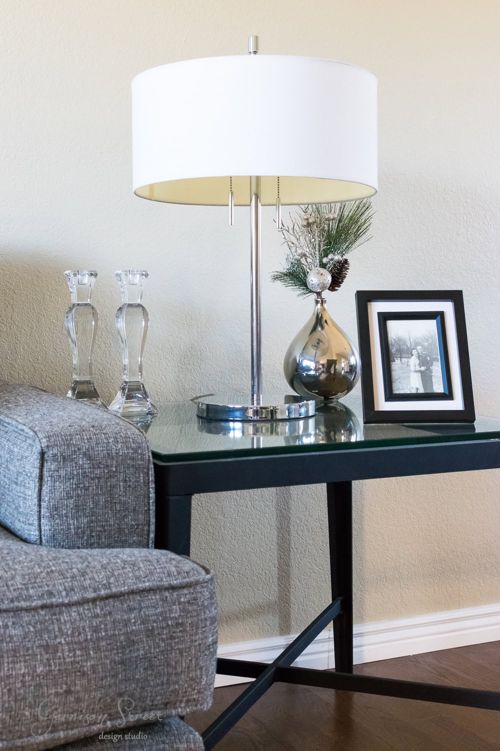 A modern living room corner features a cozy gray sofa, a black side table, and a white lamp. The table is adorned with a festive silver vase of pine branches, evoking Christmas charm, alongside a framed black and white photo and two decorative glass candlesticks.