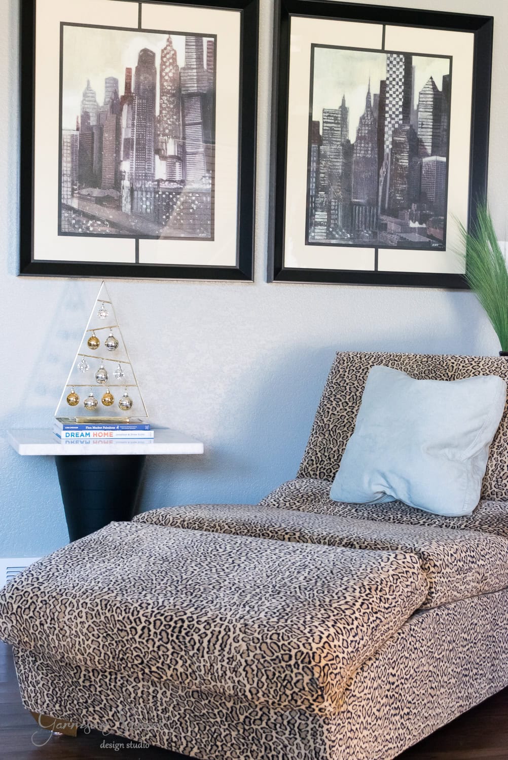 A modern room corner features a chic leopard-print lounge chair adorned with a white pillow, next to a small round table. The table displays a stack of books and a triangular decorative piece, while two framed cityscape artworks enhance the light gray wall.