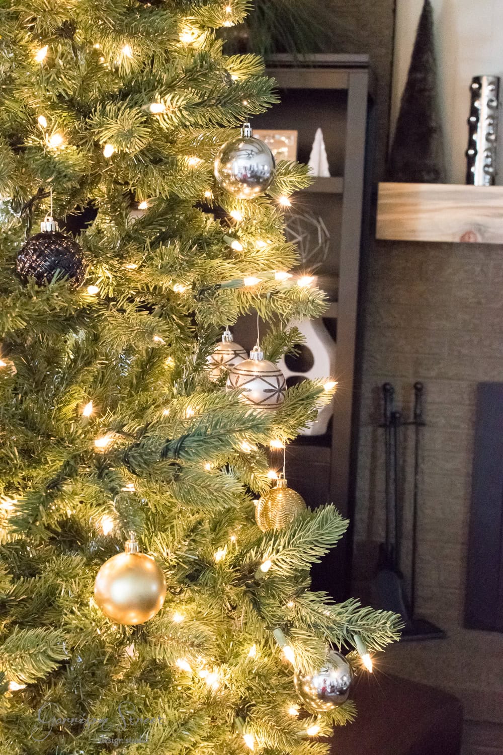 A modern Christmas tree adorned with gold, silver, and black baubles glows softly under white lights. In the background, a shelf showcases chic decorative items beside a fireplace with a sleek wooden mantel.