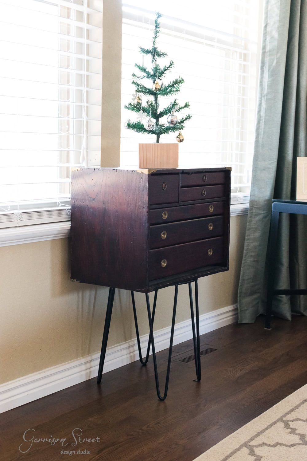 A small, modern Christmas tree in a wooden box sits atop a dark wooden chest with hairpin legs. The chest, boasting six sleek drawers, is positioned against a light-colored wall in a room with tall windows and elegant green curtains.