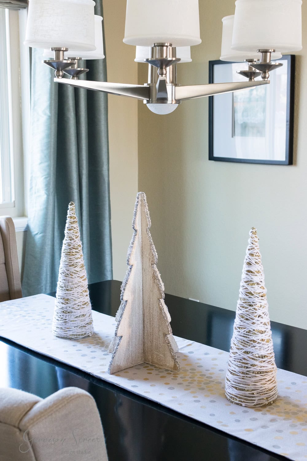 A modern dining table with a white runner showcases three decorative Christmas trees made of wood and twine. Above, a sleek chandelier with white shades hangs elegantly. In the background, blue curtains frame a window alongside a framed picture, completing the festive look.